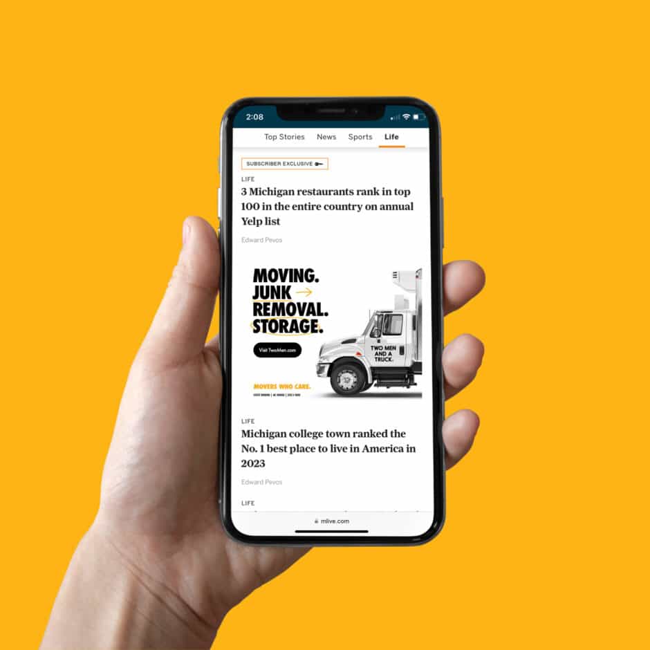 A hand holds a smartphone displaying a news article about Michigan restaurants and colleges, with an ad for moving and storage services; bright yellow background.