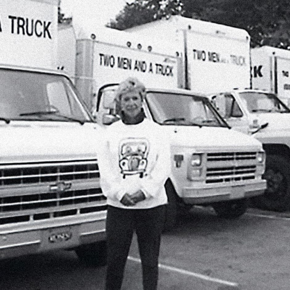 A person stands smiling in front of a row of moving trucks labeled "Two Men and a Truck." Wearing a branded sweatshirt, the scene conveys professionalism and pride.