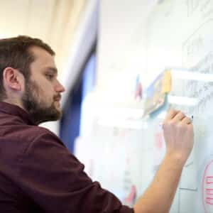 A man in a maroon shirt writes on a glass board filled with notes and diagrams. The atmosphere is focused and analytical.