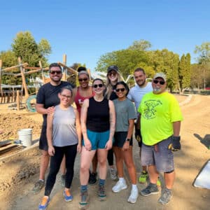 A diverse group of eight people, smiling and wearing casual attire, stand together at a sunny outdoor construction site, conveying teamwork and positivity.