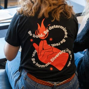 A person sits wearing a black T-shirt with a red hand and flame illustration on the back. Surrounding text reads: "Be bold. Be determined. Be real. Be human. Diversity in all we do." The overall tone is empowering and inclusive.