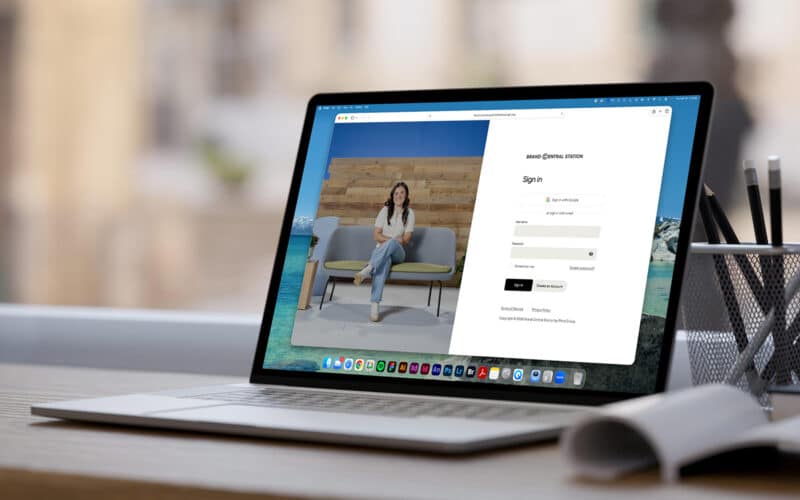 A laptop on a desk displays a sign-in page with a split screen. The left shows a woman seated on a gray couch, exuding a relaxed and professional vibe.