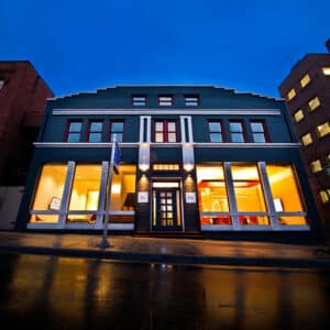 Two-story brick building at dusk with illuminated large windows revealing a warmly lit interior. Wet pavement reflects the blue evening sky.