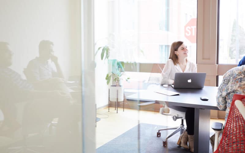 A woman smiling at a laptop sits in a bright office. Reflections of two men are seen in the glass wall, and a plant is in the corner. A stop sign is visible outside.