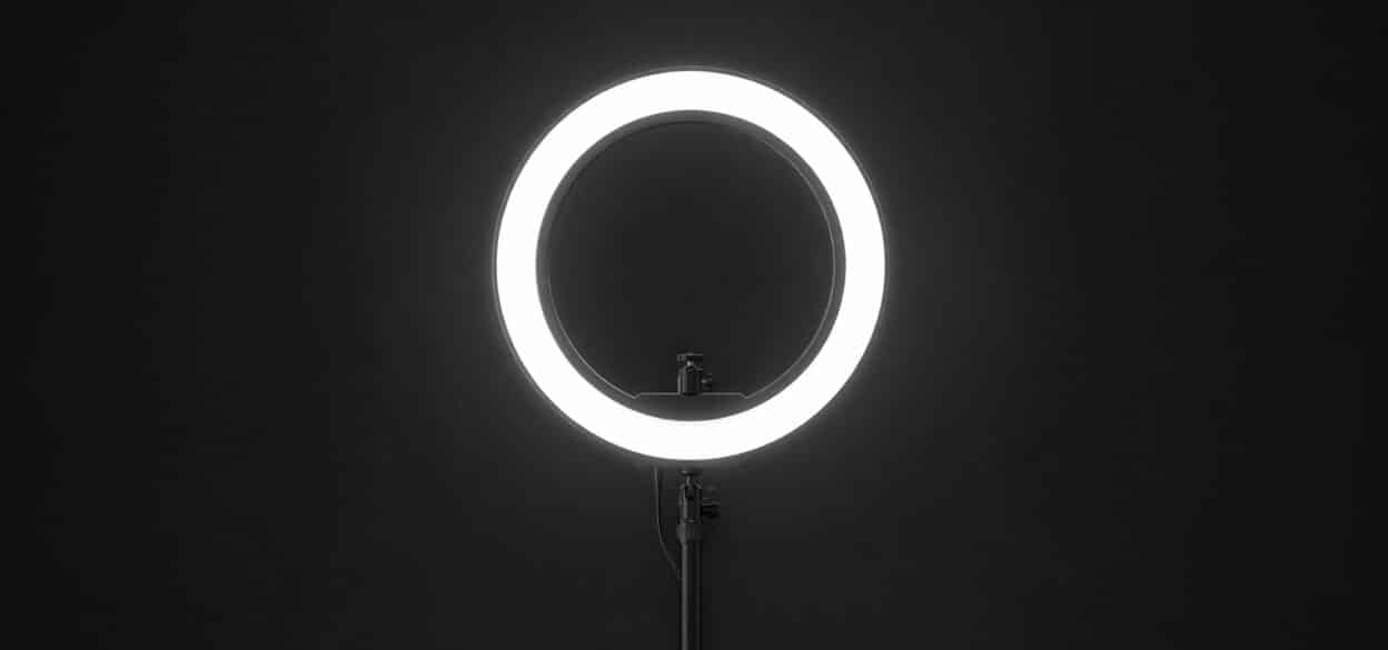 A bright circular ring light stands against a dark background, casting a soft glow. The minimalist setup conveys a clean and modern atmosphere.