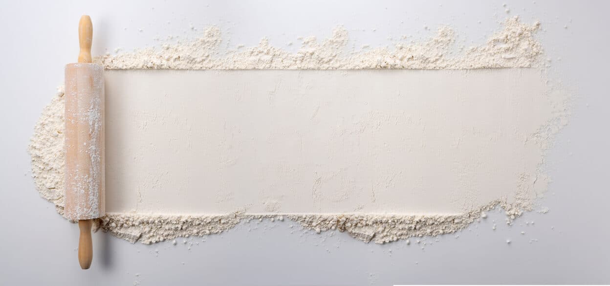 A wooden rolling pin dusted with flour rests on a white surface. Flour is scattered around, creating a border with a smooth, rolled-out center. The image conveys preparedness for baking.