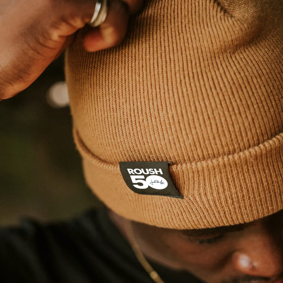Close-up of a person adjusting a tan beanie with a “ROUSH 50” label. The background is soft-focused, creating a relaxed and warm atmosphere.