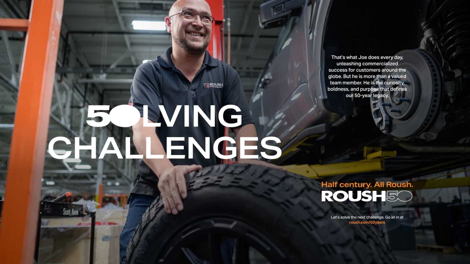 A smiling man in a workshop holds a tire near a raised vehicle. Bold text reads "Solving Challenges." Industrial background with a focus on innovation.