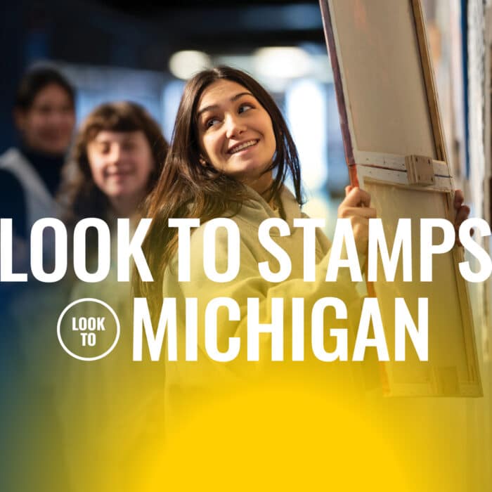 A young woman smiles while holding a large canvas in an art studio. The text reads "Look to Stamps, Michigan" in bold. The mood is creative and inspiring.