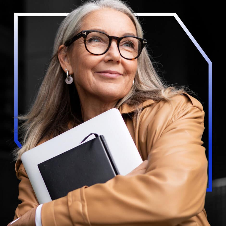 A confident woman with long gray hair and glasses, wearing a tan coat, holds a laptop and notebook. She smiles softly, conveying a sense of wisdom.