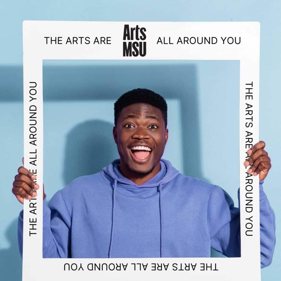 Man in a blue hoodie smiling joyfully, holding a white frame against a light blue background. The white frame says "The Arts Are All Around You" with the Arts MSU logo.