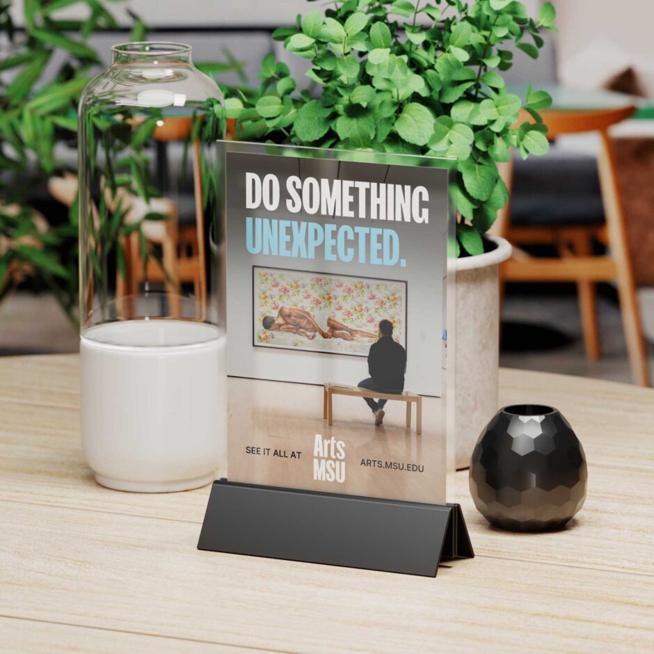 A sign on a wooden table reads "Do Something Unexpected" with an art gallery photo in the background. Tables, chairs, and plants are blurry in the background behind the sign.