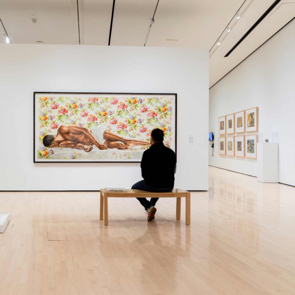 A person sits on a bench in a gallery, facing a large painting of a reclining figure against a floral background. The space is bright.