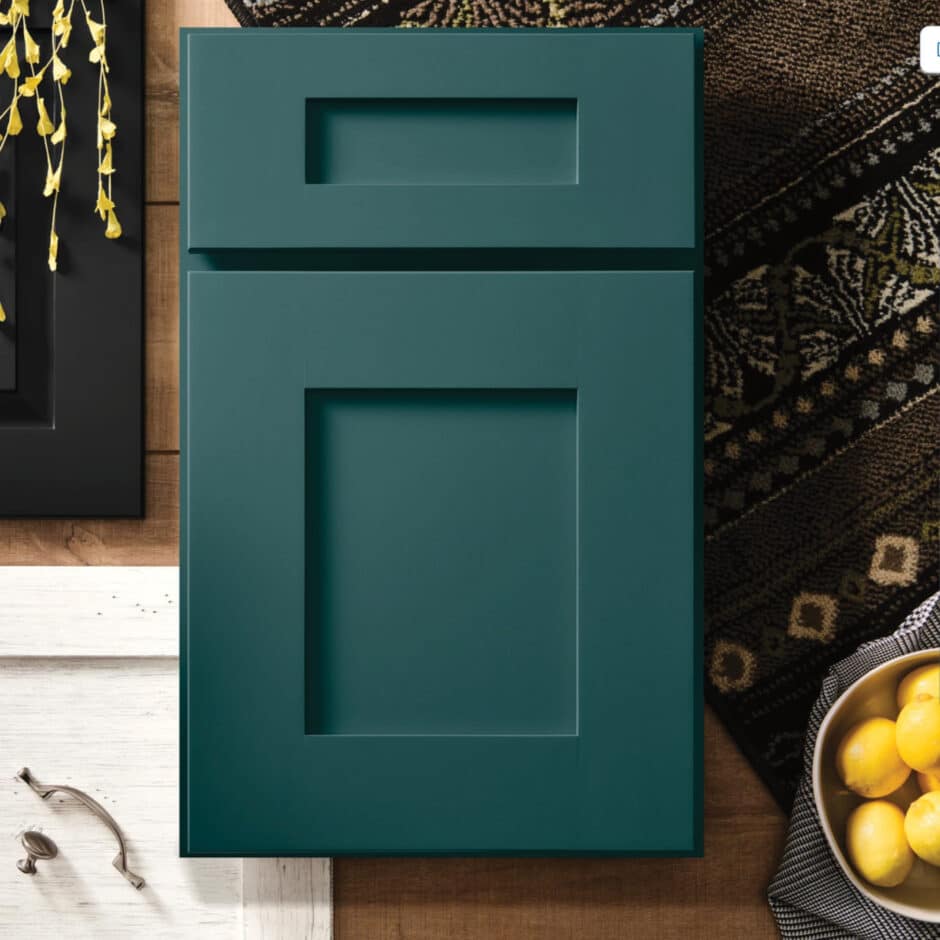 Dark teal cabinet door on a wood surface, next to patterned fabric and a bowl of lemons. Yellow flowers are visible, adding a fresh contrast.