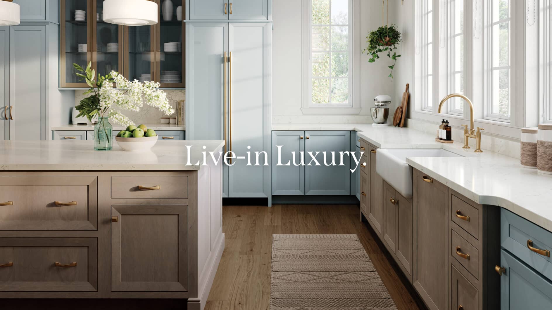 Bright, upscale kitchen with soft blue and natural wood cabinetry, white quartz countertops, a farmhouse sink, brass hardware, large windows, and a central island decorated with white flowers and a bowl of green apples. The text “Live-in Luxury.” appears over the image.
