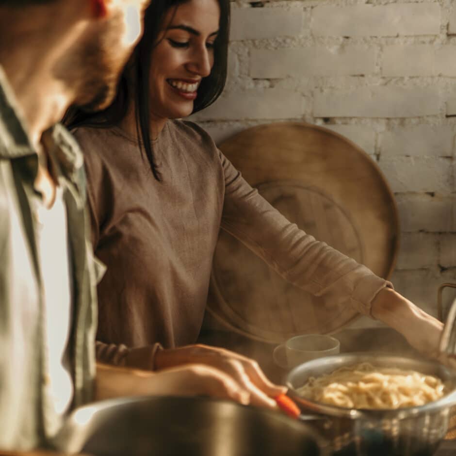 A woman in a cozy kitchen drains steaming pasta in a colander, smiling warmly at a man beside her. The scene conveys a joyful, intimate moment.