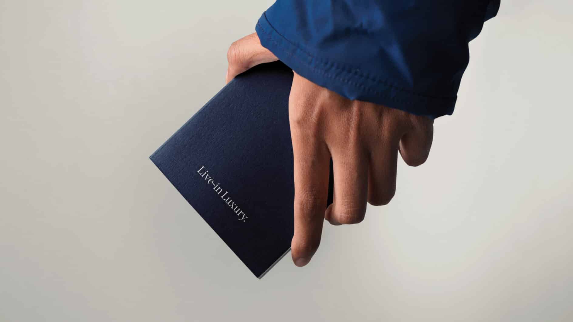 A hand in a blue sleeve holds a dark textured book with the words "Live in Luxury" in white. The tone is elegant and minimalist.