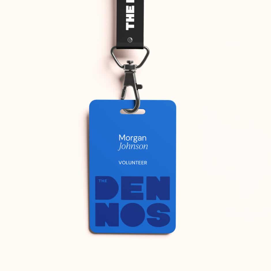 Blue volunteer badge reading “Morgan Johnson” hangs from a black lanyard and metal clip, with bold “THE DENNOS” lettering and a clean tone.