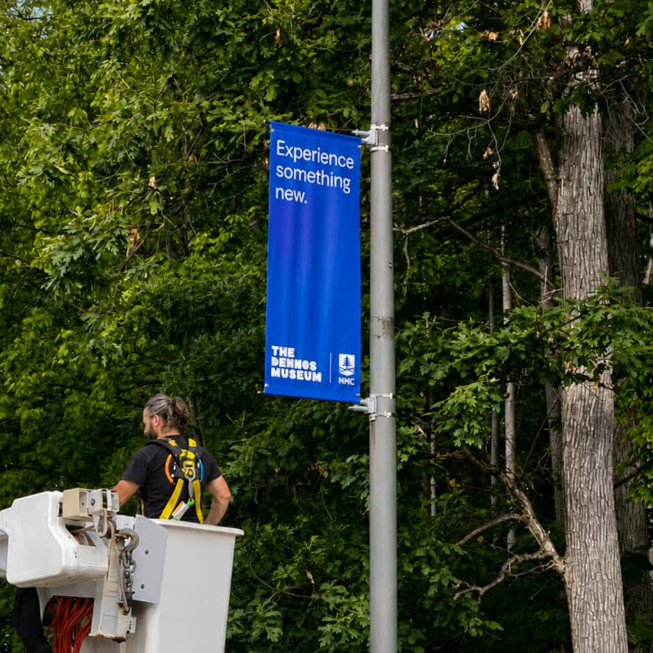 Blue “Experience something new” banner for The Demos Museum hangs beside a worker in a lift, surrounded by lush green trees.