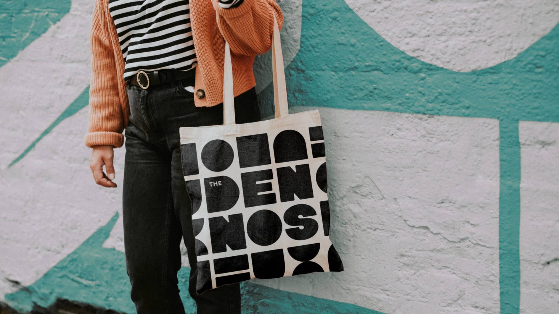 Person in an orange cardigan and striped shirt carries a bold black-and-white typography tote bag before a turquoise and white mural.