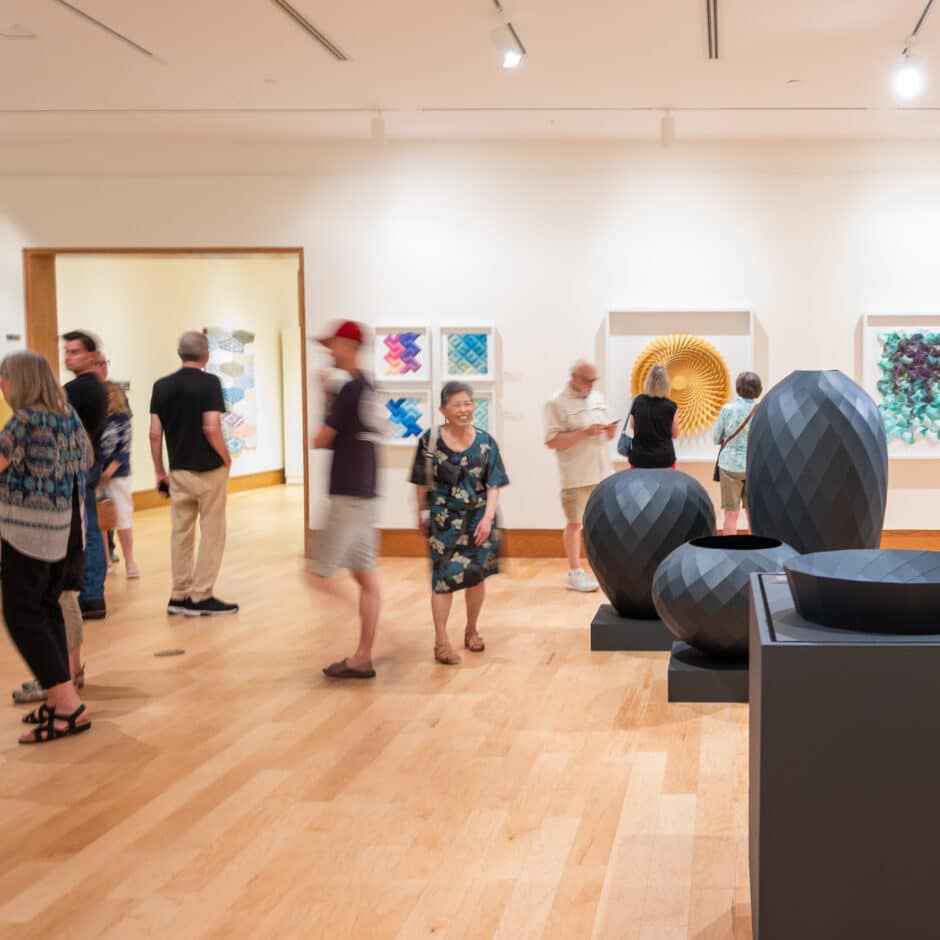 Visitors browse colorful abstract artwork and large faceted black sculptures in a bright contemporary gallery, creating a calm, lively atmosphere.