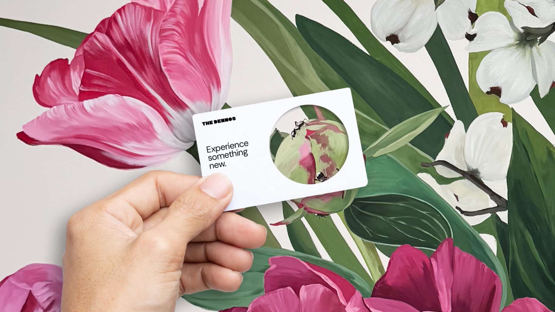 Hand holding a white card reading “Experience something new” against a vibrant botanical backdrop of pink tulips, white flowers, and green leaves.