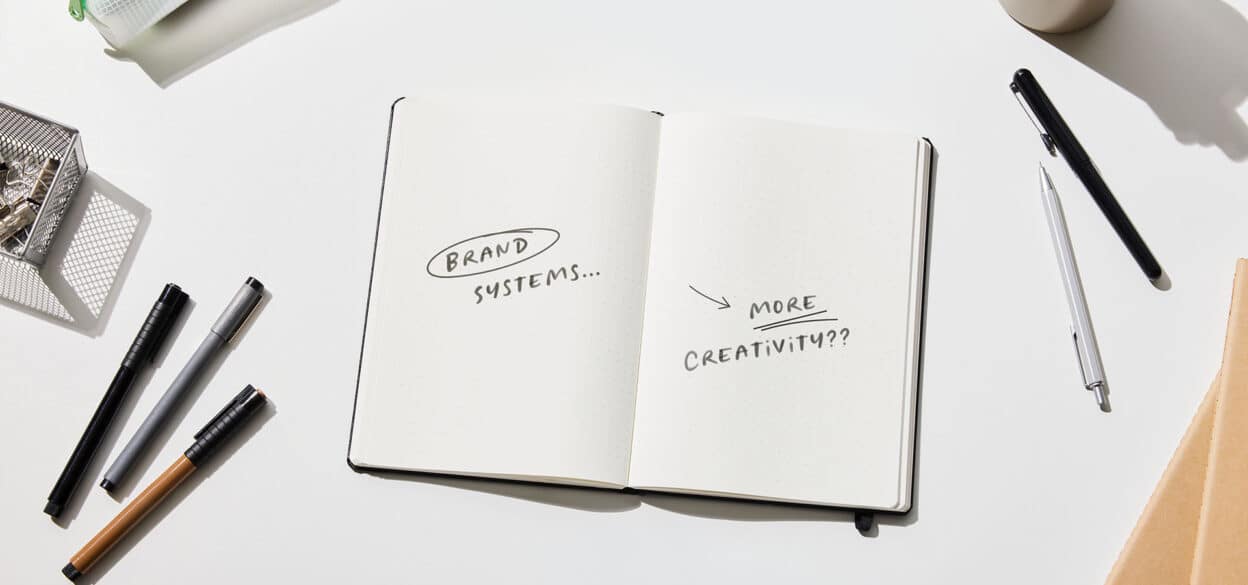 Open notebook on a white desk reads “Brand systems…” and “More creativity???” surrounded by pens, suggesting playful creative planning.