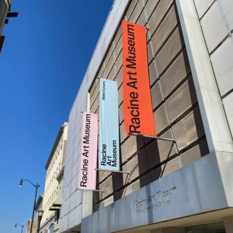 Street view of the Racine Art Museum, featuring pink, white, and orange vertical signs on a modern glass building under a clear blue sky.