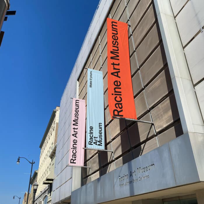 Street view of the Racine Art Museum, featuring pink, white, and orange vertical signs on a modern glass building under a clear blue sky.