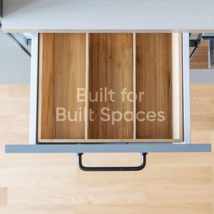 Top-down view of a modern wooden cabinet drawer with three compartments, black handle, and “Built for Built Spaces” text; clean, warm design.